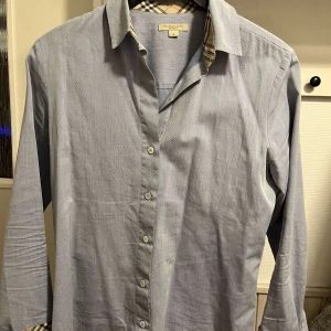Blå skjorta från Burberry - Snygg blå skjorta från Burberry med klassiskt rutigt mönster på insidan av kragen. Skjortan har långa ärmar och knappar framtill. Perfekt för en stilren look.
