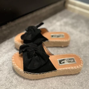 Svarta sandaler med rosett - Snygga svarta sandaler med en stor rosett på ovansidan. De har en flätad sula som ger en somrig känsla. Perfekta för varma dagar och avslappnade tillfällen.