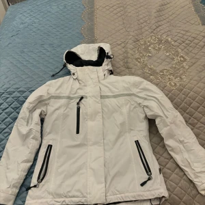 Vit jacka från Best in Town - Säljer en vit jacka från Best in Town med huva och flera dragkedjefickor. Jackan har en sportig design med svarta detaljer och är perfekt för kyligare väder.