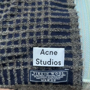 Rutig halsduk från Acne Studios - Snygg rutig halsduk från Acne Studios i en mix av grått och blått. Tillverkad i 90% virgin ull och 10% polyamid, vilket gör den både varm och hållbar. Perfekt för att hålla stilen under kyliga dagar.