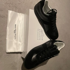 Svarta sneakers från Maison Margiela - Stilrena svarta sneakers från Maison Margiela med snörning och vit insida. Skorna har en elegant design med en kombination av skinn och syntetmaterial. Perfekta för en modern och minimalistisk look.