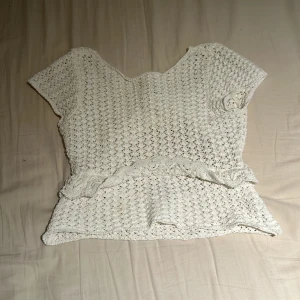 Vit stickad topp med volangdetaljer - Charmig vit stickad topp med korta ärmar och volangdetaljer. Toppen har en unik design med öppen rygg och är perfekt för en somrig look.