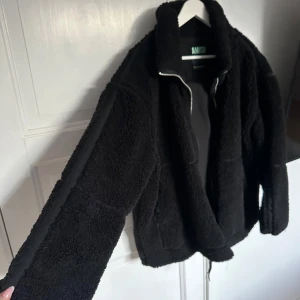 Svart teddyjacka från New Yorker  - Mysig svart teddyjacka från New Yorker med dragkedja framtill. Jackan har en avslappnad passform och långa ärmar, perfekt för kyliga dagar. Den fluffiga texturen ger en trendig look.