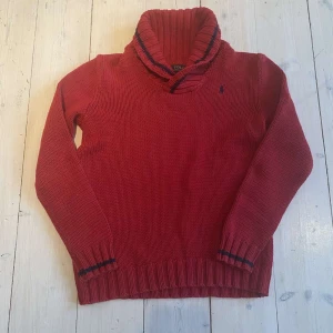 Röd stickad tröja från Ralph Lauren - Säljer en snygg röd stickad tröja från Ralph Lauren med broderad logga på bröstet. Tröjan har en hög krage och ribbade muddar vid ärmslut och nederkant. Perfekt för kyliga dagar!