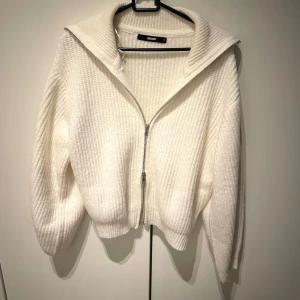 Vit ribbad kofta från Bik Bok - Säljer en stilren vit kofta från Bik Bok i storlek S. Den är ribbad med dragkedja framtill och har en hög krage. Perfekt för kyliga dagar!