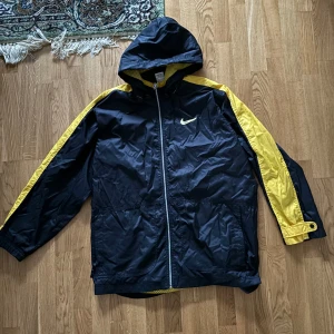 Svart och gul vindjacka från Nike - Snygg svart och gul vindjacka från Nike med huva och dragkedja. Jackan har långa ärmar och ett stort Nike-logotyp på ryggen. Perfekt för blåsiga dagar.