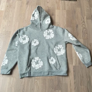 Grå hoodie från Denim Tears - Säljer en grå hoodie från Denim Tears med ett unikt vitt blommönster. Tröjan har en bekväm passform med långa ärmar och en praktisk magficka. Perfekt för en avslappnad stil.