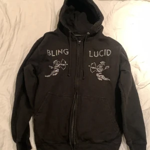 Svart blinglucid zip - Snygg svart hoodie med dragkedja och huva från Bling Lucid. Framsidan pryds av strassdetaljer i form av texten 'BLING LUCID' och två änglar. Perfekt för en cool och avslappnad stil. Storlek xs! Väldigt bra skick och inga defekter. Tveka inte att höra av er!