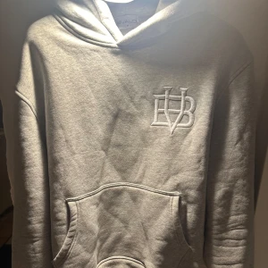 Grå hoodie från BVALDI - Säljer en stilren grå hoodie från BVALDI med en broderad logga på bröstet. Tröjan har en klassisk känguruficka och en bekväm huva. Perfekt för en avslappnad look.
