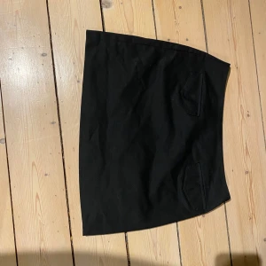 Svart kjol med fickor - Snygg svart kjol med två dekorativa fickor framtill. Kjolen har en klassisk design och är perfekt för en stilren look. Passar bra till olika tillfällen och kan enkelt matchas med olika toppar.