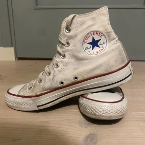 Vita Converse All Star sneakers - Klassiska vita Converse All Star sneakers med hög snörning och den ikoniska logotypen på sidan. Skorna har en röd rand längs sulan och är tillverkade i textilmaterial.