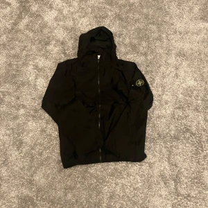 Svart jacka från Stone Island - Snygg svart jacka från Stone Island med huva och dragkedja. Den har en ikonisk logotyp på ärmen och är perfekt för en stilren look. Jackan är tillverkad i ett slitstarkt material och har en modern design. Är inte säker om den är äkta eftersom jag köpte den på loppis men den är helt ny och har inga tecken på användning.