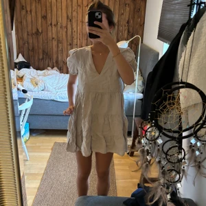 Beige kortklänning med volangdetaljer - Säljer en superfin beige kortklänning med volangdetaljer. Den har en v-ringad hals och korta ärmar som ger en söt och somrig look. Perfekt för en avslappnad dag eller en sommarfest! 💛