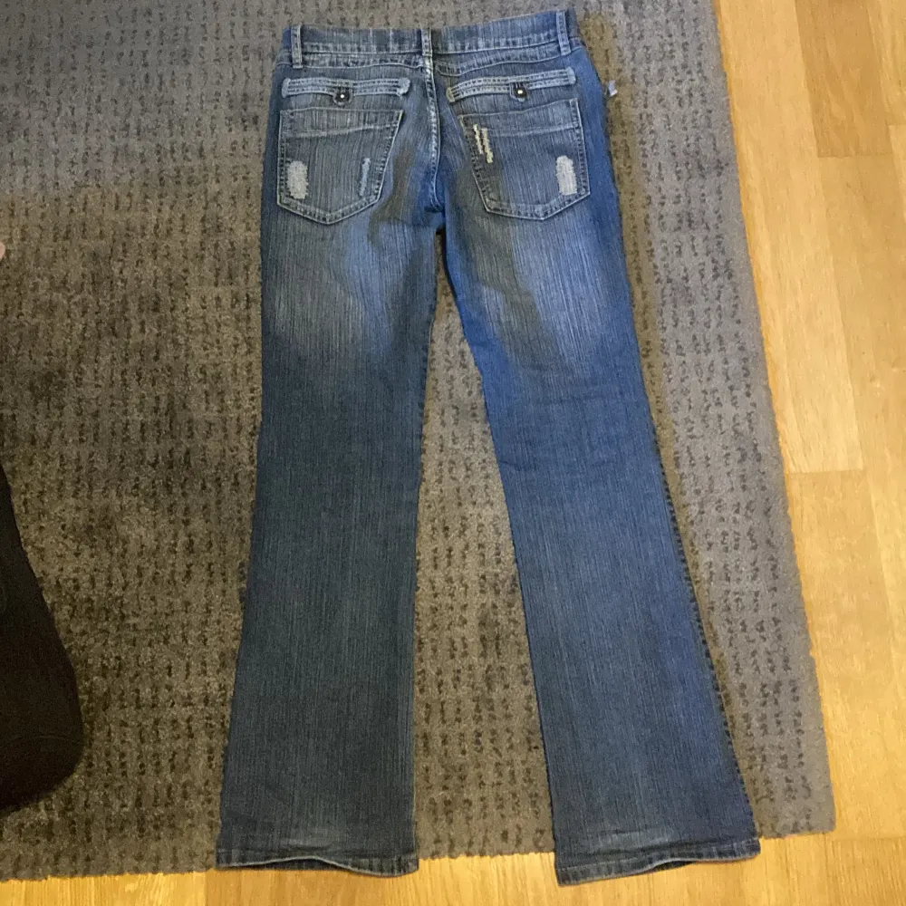 Säljer ett par blå jeans med coolt tryck på framsidan. De är lågmidjade och har en bootcut still. Jeansen har slitna detaljer på bakfickorna och är perfekta för en avslappnad stil.. Jeans & Byxor.