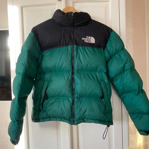  pufferjacka från The North Face - Säljer en snygg och varm pufferjacka från The North Face i grönt och svart. Jackan har en dragkedja framtill och är perfekt för kalla vinterdagar. Den är i mycket bra skick och har en klassisk stil som passar till det mesta. Perfekt för både vardag och äventyr! 