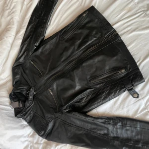 Svart skinnjacka från AMN - Säljer en snygg svart skinnjacka från AMN i mycket bra skick. Jackan har flera dragkedjor och en cool design som passar perfekt för höst och vår. Den är långärmad och har en stilren look som funkar både till vardags och fest. Perfekt för dig som vill ha en tidlös stil! 🖤