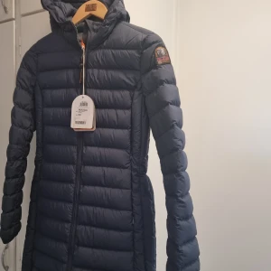 Mörkblå dunjacka från Parajumpers - Säljer en snygg och varm mörkblå dunjacka från Parajumpers. Jackan har en lång passform med dragkedja och en mysig huva. Perfekt för kalla vinterdagar! Den är i nytt skick med etikett kvar. Passar perfekt för både vardag och vinteräventyr. Original  pris 5999 kr