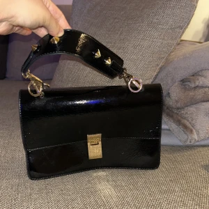 Svart axelväska från Gina Tricot - Snygg svart axelväska i skinn från Gina Tricot med coola nitar på axelremmen. Väskan har en stilren design med ett guldfärgat spänne framtill. Perfekt för en utekväll eller fest! 🖤✨