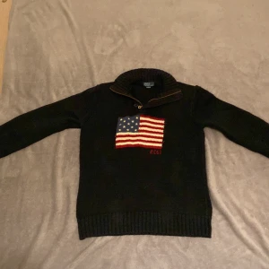 Svart stickad half zip tröja från Polo Ralph Lauren - Säljer en snygg svart stickad tröja från Polo Ralph Lauren med en cool amerikansk flagga på framsidan och RL67 broderat. Tröjan har en dragkedja vid halsen och är perfekt för höst och vinter. Den är i mycket bra skick och supermysig att ha på sig!
