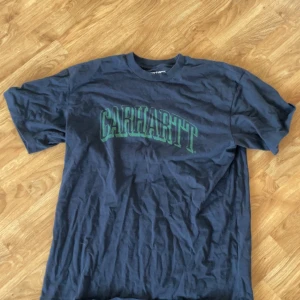 Blå t-shirt från Carhartt - Säljer en snygg blå t-shirt från Carhartt i storlek M. Den har ett coolt grönt tryck med Carhartt-loggan på framsidan. Perfekt för vardagsbruk eller avslappnade tillfällen. T-shirten är i bra skick och gjord av mjuk bomull.