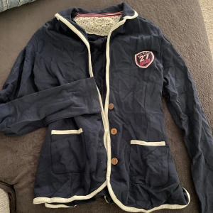 Mörkblå jacka från Hampton Republic - Säljer en snygg mörkblå jacka från Hampton Republic med vita kanter och knappar. Den har ett coolt emblem på bröstet och texten 'Hampton Republic' på ärmen. Perfekt för en avslappnad stil. Passar bra till både vardag och fest!