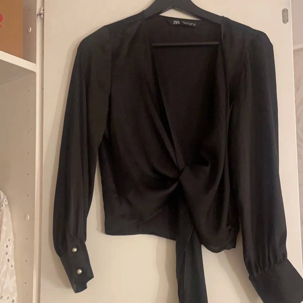 Snygg svart omlottopp från Zara i ett glansigt material. Toppen har långa ärmar med knappar vid ärmsluten och en knytning framtill som ger en elegant look. Perfekt för festliga tillfällen eller en utekväll. Passar bra till både jeans och kjol!. Toppar.