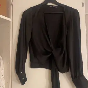 Snygg svart omlottopp från Zara i ett glansigt material. Toppen har långa ärmar med knappar vid ärmsluten och en knytning framtill som ger en elegant look. Perfekt för festliga tillfällen eller en utekväll. Passar bra till både jeans och kjol!