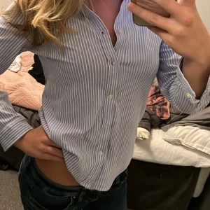 Randig skjorta från Ralph Lauren dam - Säljer en snygg blå- och vitrandig skjorta från Ralph Lauren i storlek XL. Skjortan har långa ärmar och klassiska knappar framtill. Den är i mycket bra skick och perfekt för både vardag och fest. Den broderade loggan på bröstet ger en extra touch av stil. Passar perfekt till jeans eller chinos!