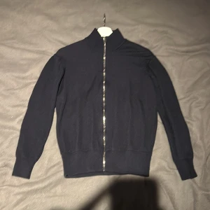 Mörkblå merinoullskofta från Asket - Säljer en stilren och bekväm mörkblå kofta från Asket i merinoull. Den har en praktisk dragkedja framtill och är perfekt för både vardag och lite finare tillfällen. Koftan är i mycket bra skick och passar perfekt för höst och vinter. Perfekt för dig som gillar en klassisk look!