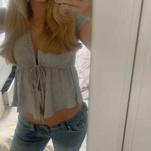 Grå kortärmad topp med knyt - Säljer en söt grå topp med korta ärmar och knytning framtill. Den har en avslappnad passform och är perfekt för sommardagar. Toppen är croppad och ger en trendig look tillsammans med jeans eller kjol.