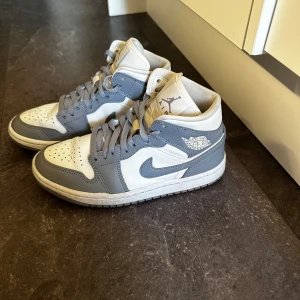 Nike Air Jordans 1 Mid - Säljer ett par Nike Air Jordans i grått och vitt. Skorna är i mycket bra skick och har en snygg design med snörning. Perfekta för både vardag och fest. Storlek dam 38🩵🩵Nypris 2800kr