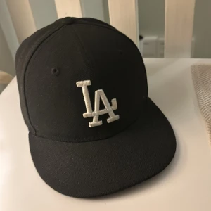 Svart keps från New Era - Säljer en svart keps från New Era med LA-logga framtill. Kepsen är i mycket bra skick och har en klassisk stil. Perfekt för både vardag och sportiga tillfällen. Storleken är 6 3/4, vilket motsvarar 53.9 cm i omkrets.