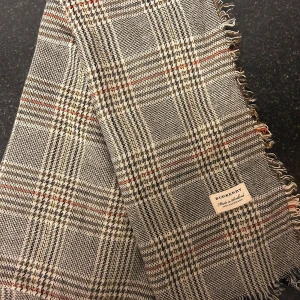 Burberry Halsduk - Säljer en klassisk rutig halsduk från Burberry i grått, beige och rött. Den är i mycket bra skick och har fransar längs kanterna. Perfekt för höst och vinter för att hålla dig varm och stilren. Märket är tydligt synligt och ger en lyxig touch.