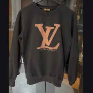 Svart tröja från Louis Vuitton - Snygg svart tröja från Louis Vuitton med det ikoniska LV-logotypen i en kontrasterande färg på framsidan. Tröjan har en klassisk rund hals och långa ärmar, perfekt för en stilren look. Står Large på lappen men sitter som en Medium.