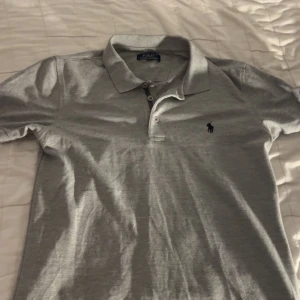 Grå pikétröja från Ralph Lauren - Säljer en stilren grå pikétröja från Ralph Lauren med klassisk krage och knappar framtill. Tröjan har en broderad logotyp på bröstet och är perfekt för en avslappnad men ändå snygg look.