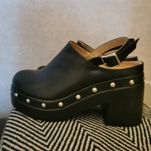 Svarta träskor  - Snygga svarta klackskor med nitar runt sulan och en justerbar rem. Perfekta för att ge en edgy touch till din outfit. Skorna har en robust klack och en stilren design. Osäker på märke, köpta i Italien. Oanvända då de inte riktigt är min stil men väldigt sköna ❤️
