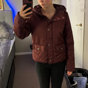 Vinröd jacka från Noisy May - Säljer en vinröd jacka från Noisy May med huva och knappar. Jackan har långa ärmar och praktiska fickor framtill. Perfekt för kyligare dagar. Den passar som en s