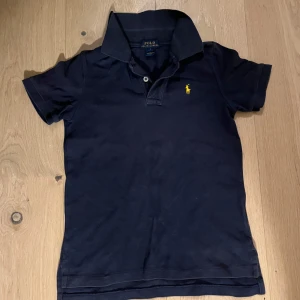 Mörkblå pikétröja från Ralph Lauren - Säljer en klassisk mörkblå pikétröja från Ralph Lauren med korta ärmar och den ikoniska gula broderade logotypen på bröstet. Perfekt för en stilren och avslappnad look. Tröjan har en krage och knappar framtill.