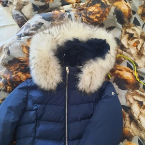 Blå dunjacka från Rockandblue - Snygg blå dunjacka från Rockandblue med pälskantad huva och dragkedja. Jackan har långa ärmar med dragkedjedetaljer vid ärmsluten. Använd ett fåtal ggr. Finns några fläckar i nack kragen men går att tvätta bort.