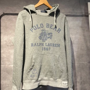Ralph Lauren hoodie - Säljer en grå hoodie från Ralph Lauren med Polo Bear-tryck och texten '1967'. Tröjan har en känguruficka och justerbar huva med snören. Perfekt för en avslappnad stil. Nypris ca 3000kr