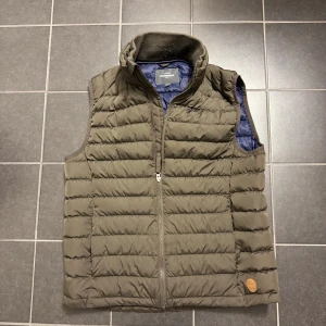 Brun quiltad dunväst - Säljer en brun quiltad dunväst med dragkedja. Perfekt för kyligare dagar och har en stilren design. Västen är ärmlös och har en liten logotypdetalj på nederkanten. Passar bra för lager-på-lager-stil.