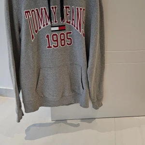 Grå hoodie från Tommy Jeans - Säljer en grå hoodie från Tommy Jeans med texten '1985' i rött och vitt framtill. Den har en känguruficka och svarta snören vid huvan. Perfekt för en avslappnad stil.
