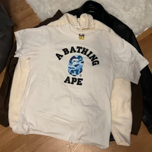 Vit A Bathing Ape T-shirt med camo-tryck - Säljer en vit A Bathing Ape T-shirt med det ikoniska camo-trycket i blått och svart. T-shirten har korta ärmar och en klassisk passform. Perfekt för dig som gillar streetwear och vill sticka ut med en unik design.