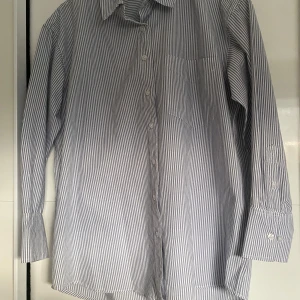 Randig skjorta - Snygg randig skjorta i blått och vitt med klassisk krage och knappar framtill. Skjortan har en bröstficka och långa ärmar med knappar vid ärmsluten. Perfekt för en stilren look. Och är oversized