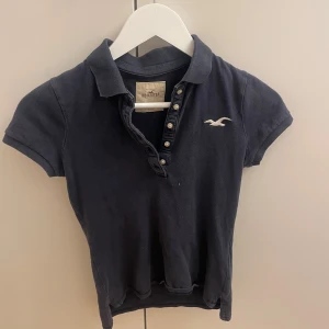 Mörkblå pikétröja från Hollister - Säljer en mörkblå pikétröja från Hollister med korta ärmar och knappar framtill. Den har en klassisk krage och Hollisters logga broderad på bröstet. Perfekt för en avslappnad stil.
