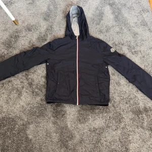 Modell urville giubottoSvart vindjacka från Moncler - Säljer en stilren svart moncler urville giubotto vindjacka med huva och dragkedja. Jackan har långa ärmar och en grå insida. Perfekt för blåsiga dagar och har en klassisk Moncler-logga på ärmen. Moddelen passar 165-180 och är xl i barn