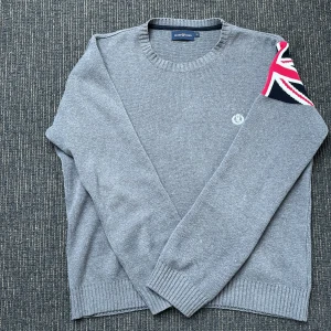 Grå tröja från Henri Lloyd  - Säljer nu en riktigt snygg grå tröja från Henri Lloyd med en unik flaggdetalj på ärmen. Tröjan har en rund halsringning och ribbade muddar. Perfekt för en stilren look.