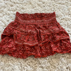 Röd volangkjol med spetsdetaljer - Säljer en superfin röd kortkjol med volanger och spetsdetaljer. Kjolen har ett elastiskt midjeband och ett vackert mönster som ger en bohemisk känsla. Perfekt för sommarens alla äventyr! 