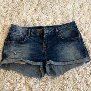 Blå jeansshorts från LTB - Snygga blå lågmidjade jeansshorts från LTB med slitna detaljer och uppvikta kanter. De har en klassisk femficksdesign och knappgylf. Perfekta för en avslappnad stil.