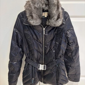 Svart vinterjacka från Michael Kors - Snygg svart vinterjacka från Michael Kors med en lyxig fuskpälsdetalj på kragen (avtagbar, se bild 3). Jackan har en quiltad design och ett bälte i midjan för en figursydd look. Den är perfekt för kyliga dagar med sin dragkedja framtill och långa ärmar. Finns även svart luva till (avtagbar). 